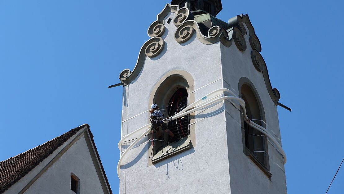 Blick auf das Glockengeschoss des Turmes der Luzerner Peterskapelle, das von weissen Rohren umschlungen ist.