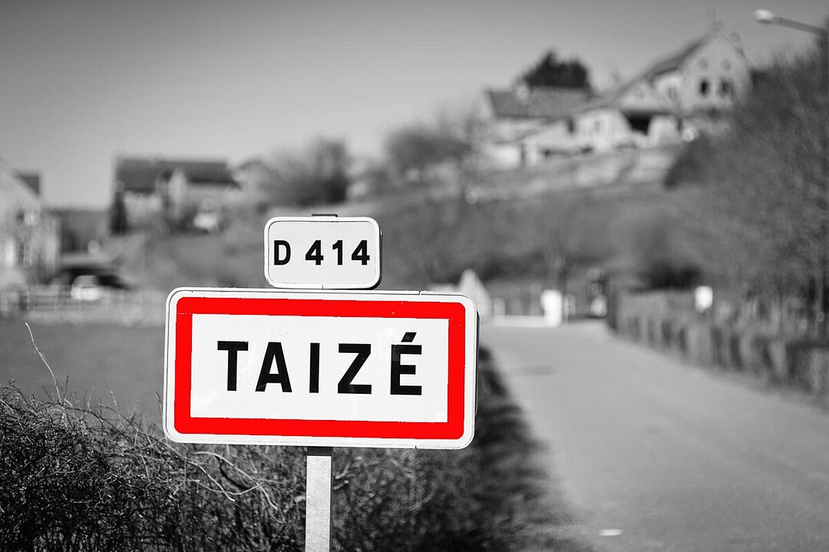 Rotumrandetes Ortsschild von Taizé vor schwarzweisser Landschaft mit Ortseingang von Taizé